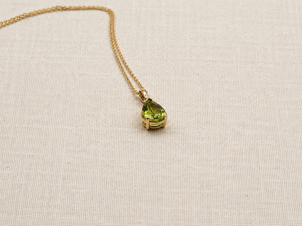 An elegant 10k solid yellow gold pendant necklace featuring a teardrop-shaped natural green peridot gemstone by Ophelisse, displayed closed up, side angle view, on a beige fabric background.