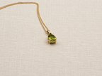 An elegant 10k solid yellow gold pendant necklace featuring a teardrop-shaped natural green peridot gemstone by Ophelisse, displayed closed up, side angle view, on a beige fabric background.