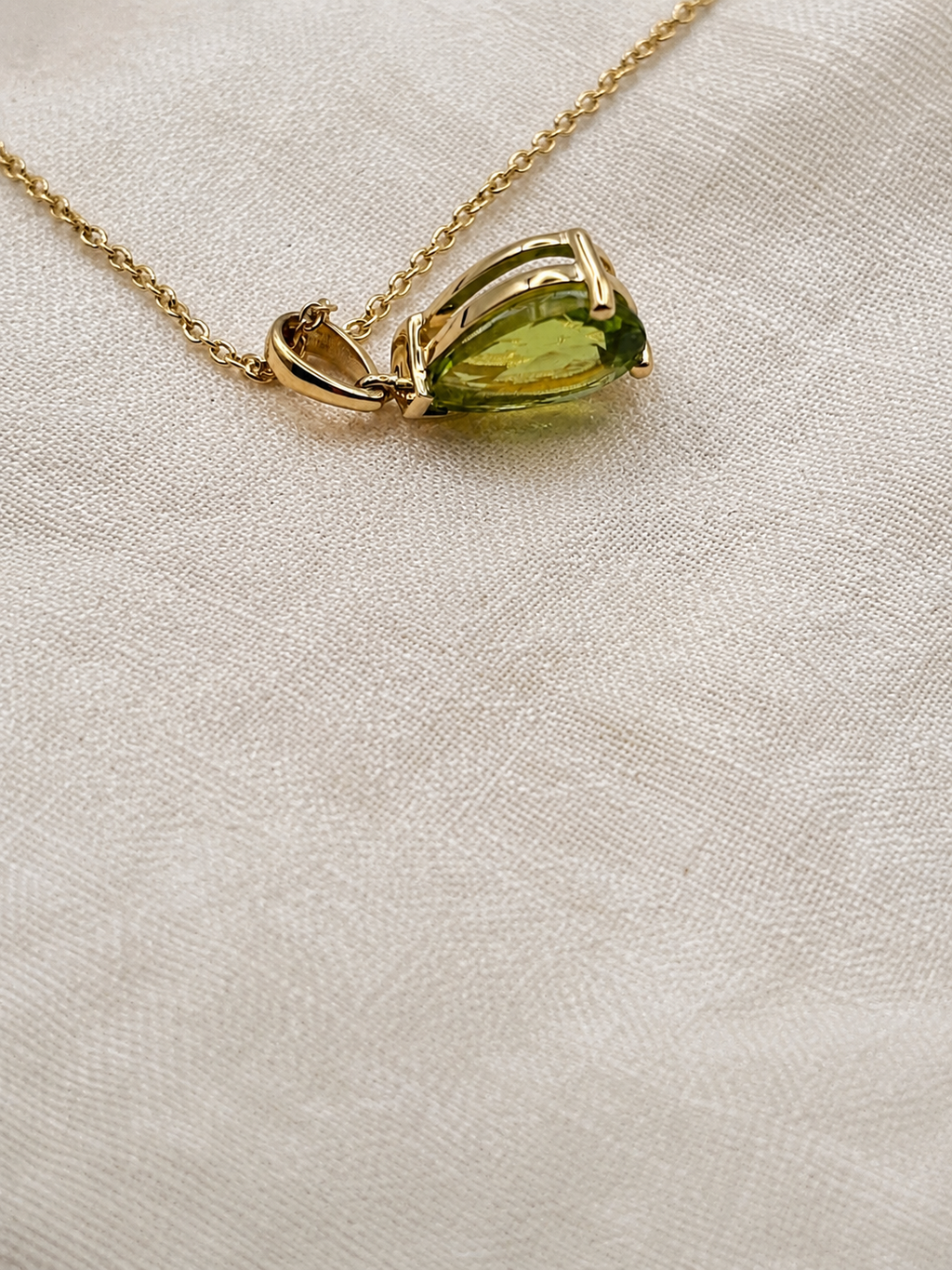 A 10k solid gold chain necklace with a teardrop-shaped natural green peridot gemstone pendant made by Ophelisse, resting on an elegant textured beige fabric background.
