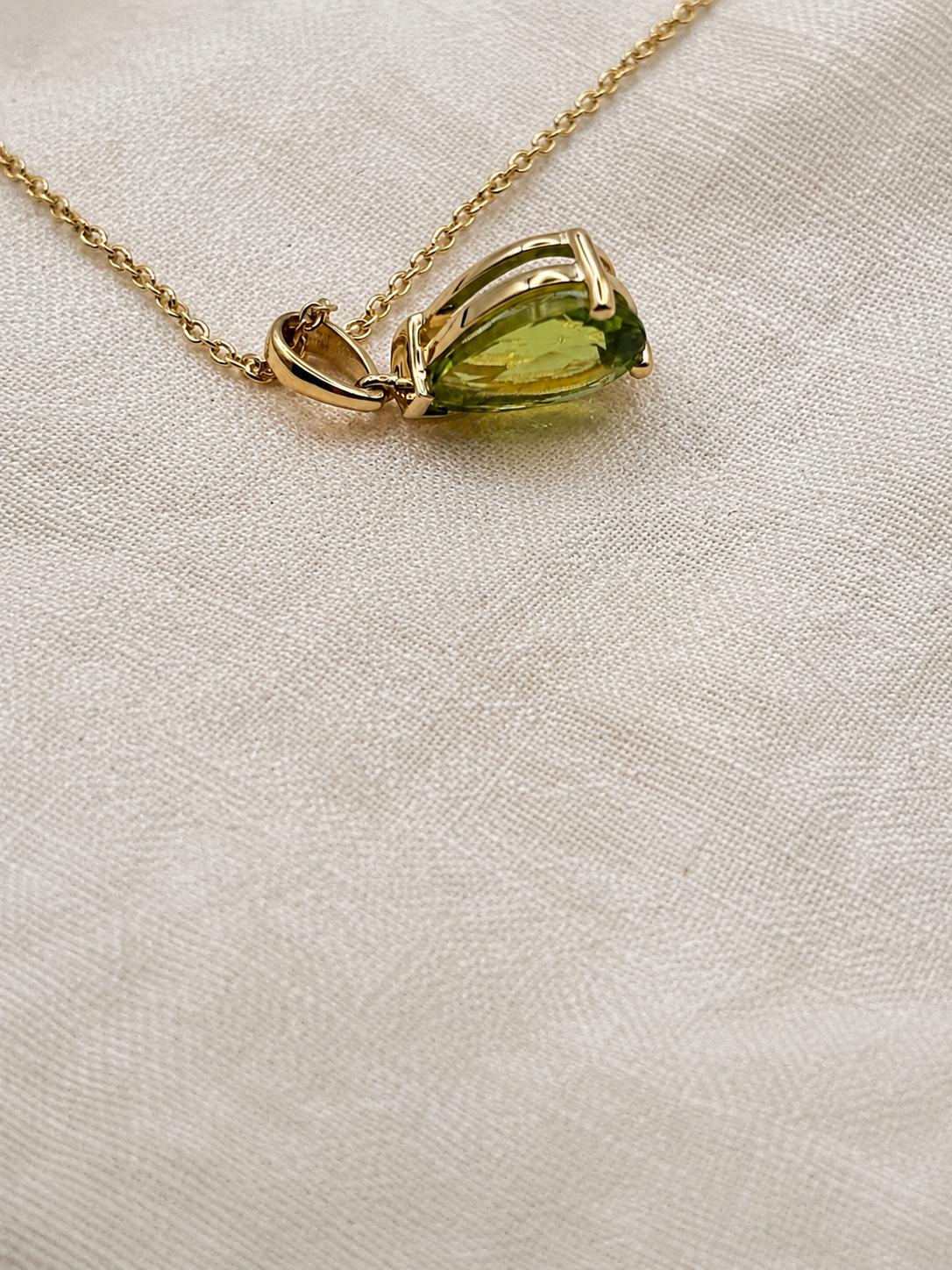 A 10k solid gold chain necklace with a teardrop-shaped natural green peridot gemstone pendant made by Ophelisse, resting on an elegant textured beige fabric background.