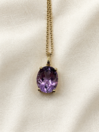An oval natural purple amethyst pendant necklace in a solid 10k yellow gold setting on a delicate gold chain by Ophelisse, displayed on a textured off-white fabric background.