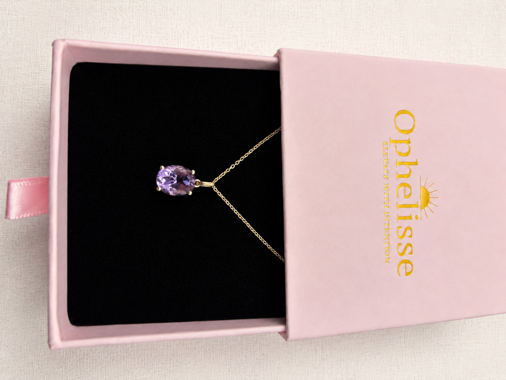 An oval-cut natural purple amethyst gemstone pendant necklace in a solid 10k yellow gold setting, displayed inside an open pink jewelry box with a black interior. The box lid is propped open, showing the gold-foiled 'Ophelisse' brand logo.