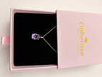 An oval-cut natural purple amethyst gemstone pendant necklace in a solid 10k yellow gold setting, displayed inside an open pink jewelry box with a black interior. The box lid is propped open, showing the gold-foiled 'Ophelisse' brand logo.