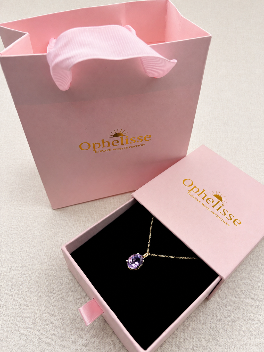 An Ophelisse pendant necklace with a purple natural amethyst gemstone on a solid 10k yellow gold chain, displayed in an open pink jewelry box next to a pink branded shopping bag and closed lid on a light fabric background.
