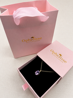 An Ophelisse pendant necklace with a purple natural amethyst gemstone on a solid 10k yellow gold chain, displayed in an open pink jewelry box next to a pink branded shopping bag and closed lid on a light fabric background.