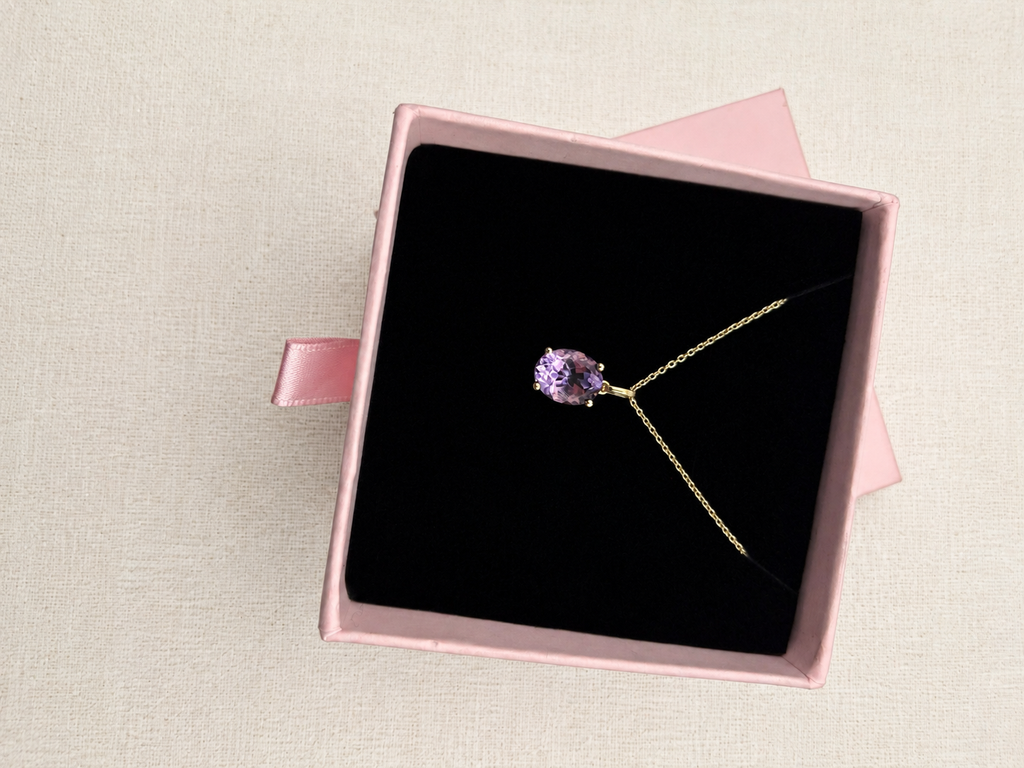 A solid 10k yellow gold necklace with a purple oval natural amethyst gemstone pendant, presented inside an open pink jewelry box with a black velvet insert by Ophelisse, set against a light beige textured background.