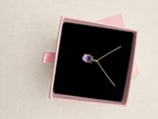 A solid 10k yellow gold necklace with a purple oval natural amethyst gemstone pendant, presented inside an open pink jewelry box with a black velvet insert by Ophelisse, set against a light beige textured background.