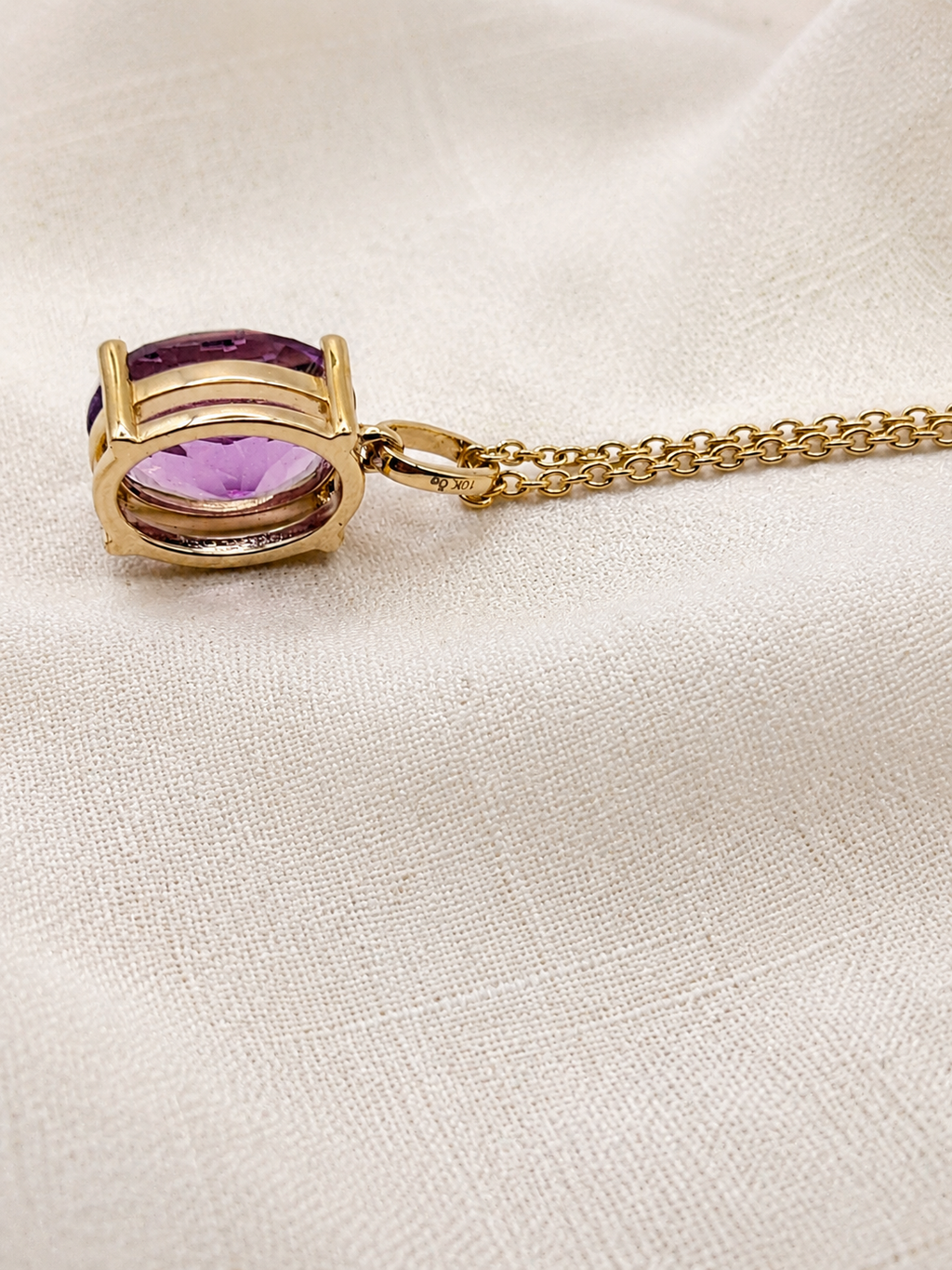 A solid 10k yellow gold pendant necklace featuring the backside of an oval natural purple amethyst gemstone by Ophelisse, resting on a textured beige linen fabric background.