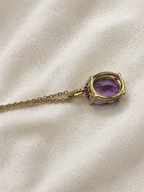 A solid 10k yellow gold pendant necklace featuring the backside of an oval natural purple amethyst gemstone by Ophelisse, resting on a textured cream-colored fabric background.