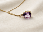A solid 10k yellow gold pendant necklace featuring an oval-cut natural purple amethyst gemstone by Ophelisse, resting on a textured cream fabric background.