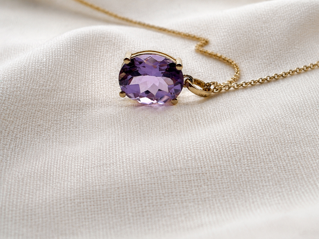 A solid 10k yellow gold pendant necklace featuring a faceted natural purple amethyst gemstone by Ophelisse, resting on a textured off-white fabric background.