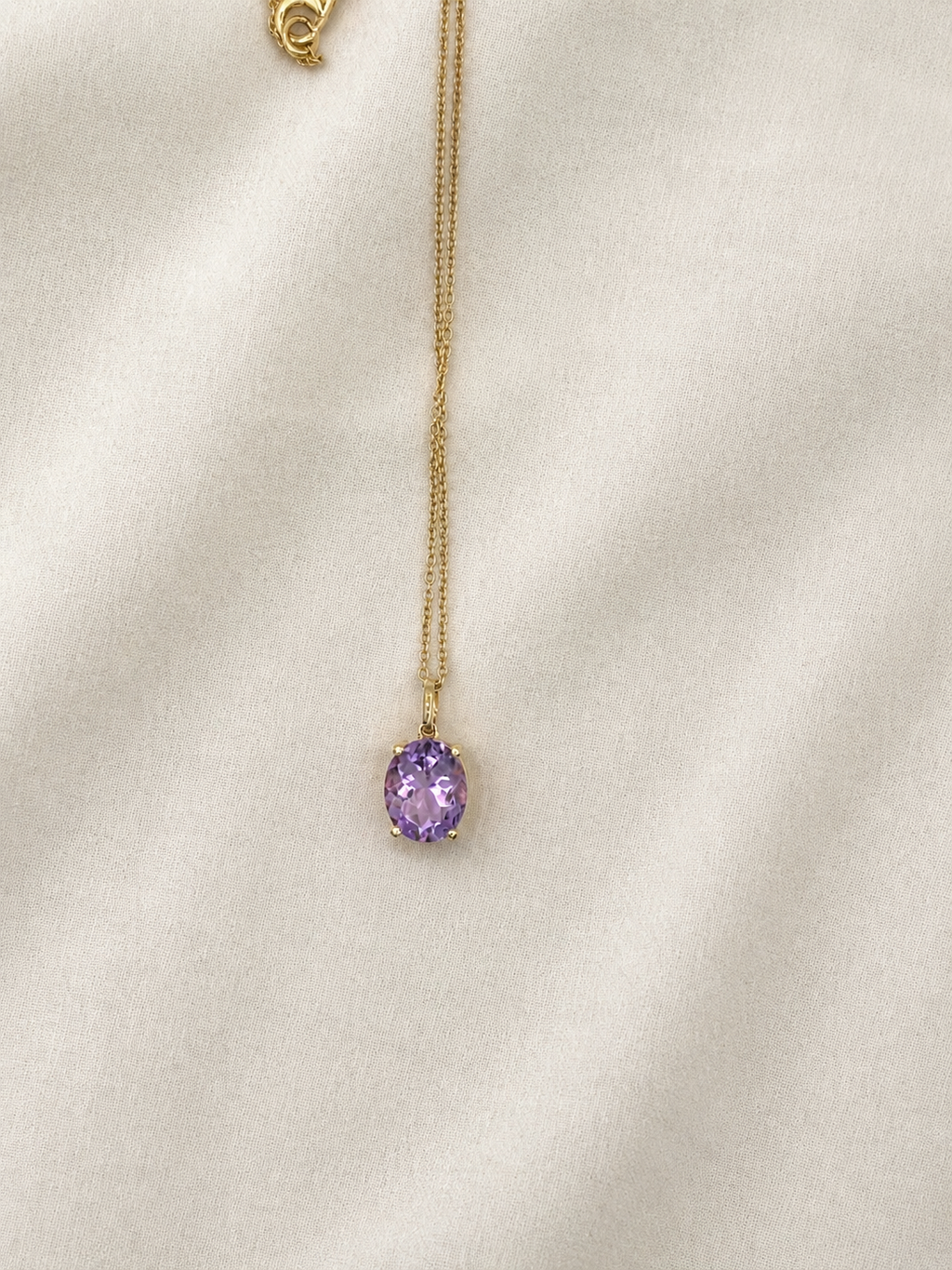 A delicate solid 10k yellow gold chain necklace with an oval natural purple amethyst gemstone pendant by Ophelisse, displayed on a soft, textured off-white fabric background.