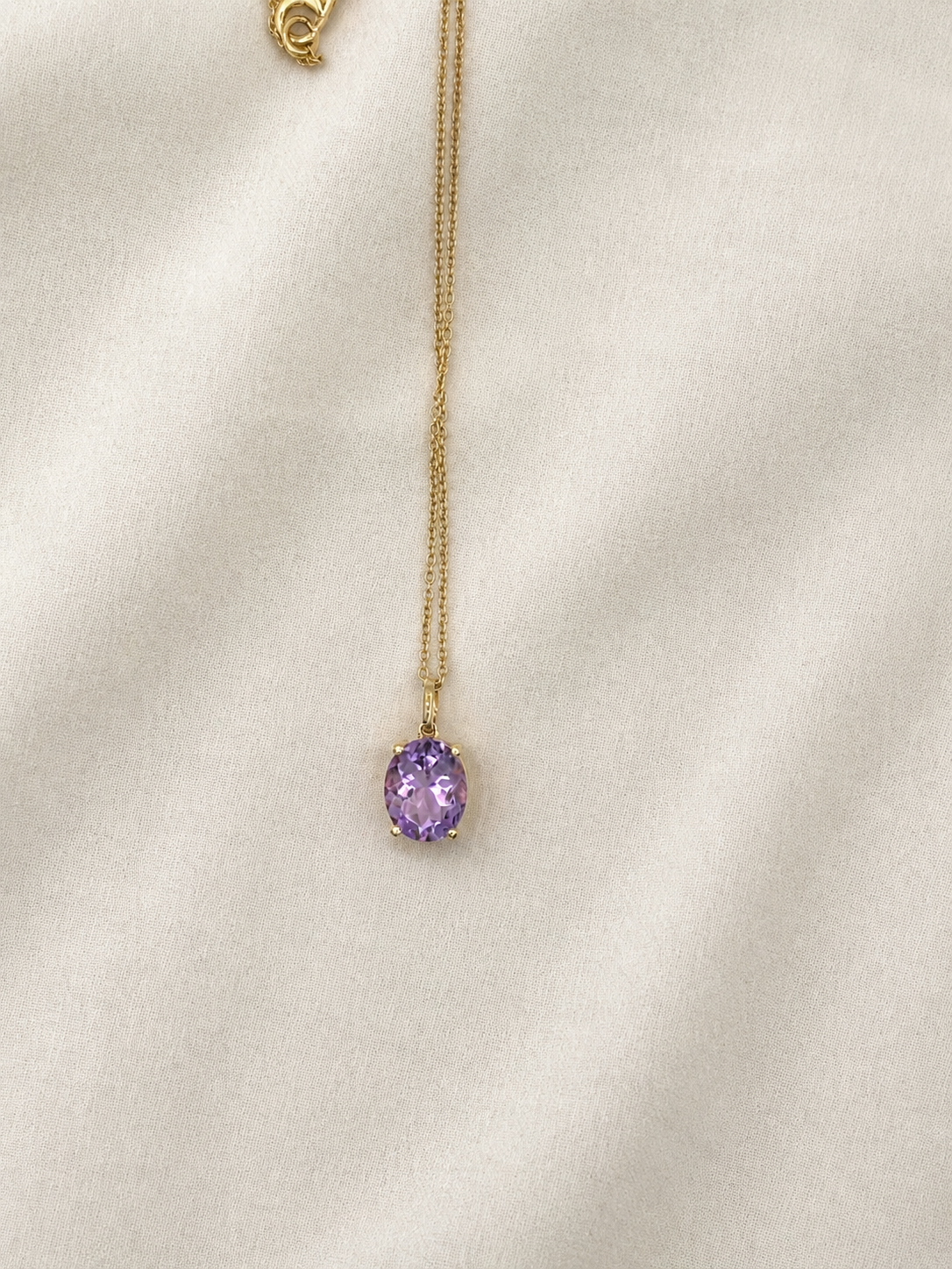 A delicate solid 10k yellow gold chain necklace with an oval natural purple amethyst gemstone pendant by Ophelisse, displayed on a soft, textured off-white fabric background.