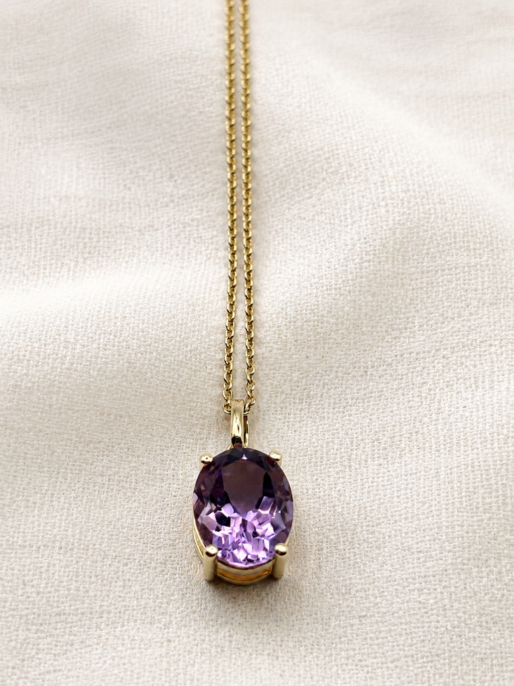 A solid 10k yellow gold chain necklace with an oval natural purple amethyst gemstone pendant by Ophelisse, displayed on a textured off-white fabric background.