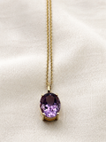 A solid 10k yellow gold chain necklace with an oval natural purple amethyst gemstone pendant by Ophelisse, displayed on a textured off-white fabric background.