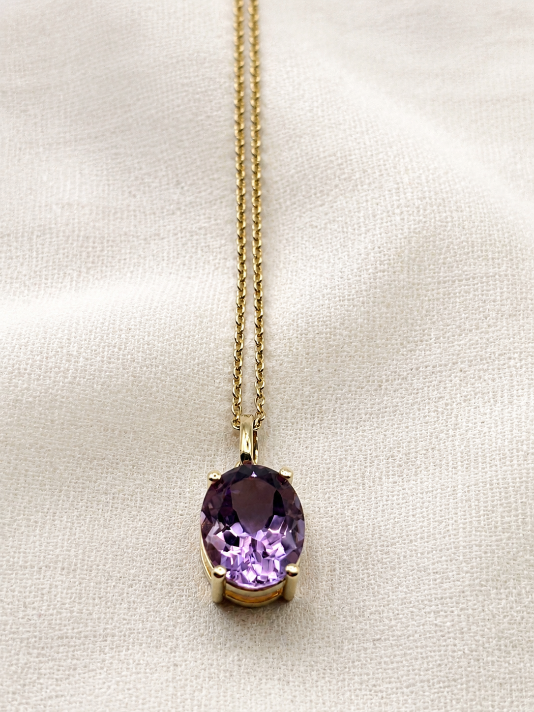 A solid 10k yellow gold chain necklace with an oval natural purple amethyst gemstone pendant by Ophelisse, displayed on a textured off-white fabric background.