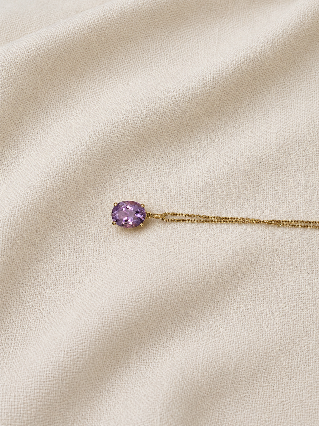 A solid 10k yellow gold chain necklace with an oval natural purple amethyst gemstone pendant by Ophelisse, displayed on a textured cream-colored fabric background.