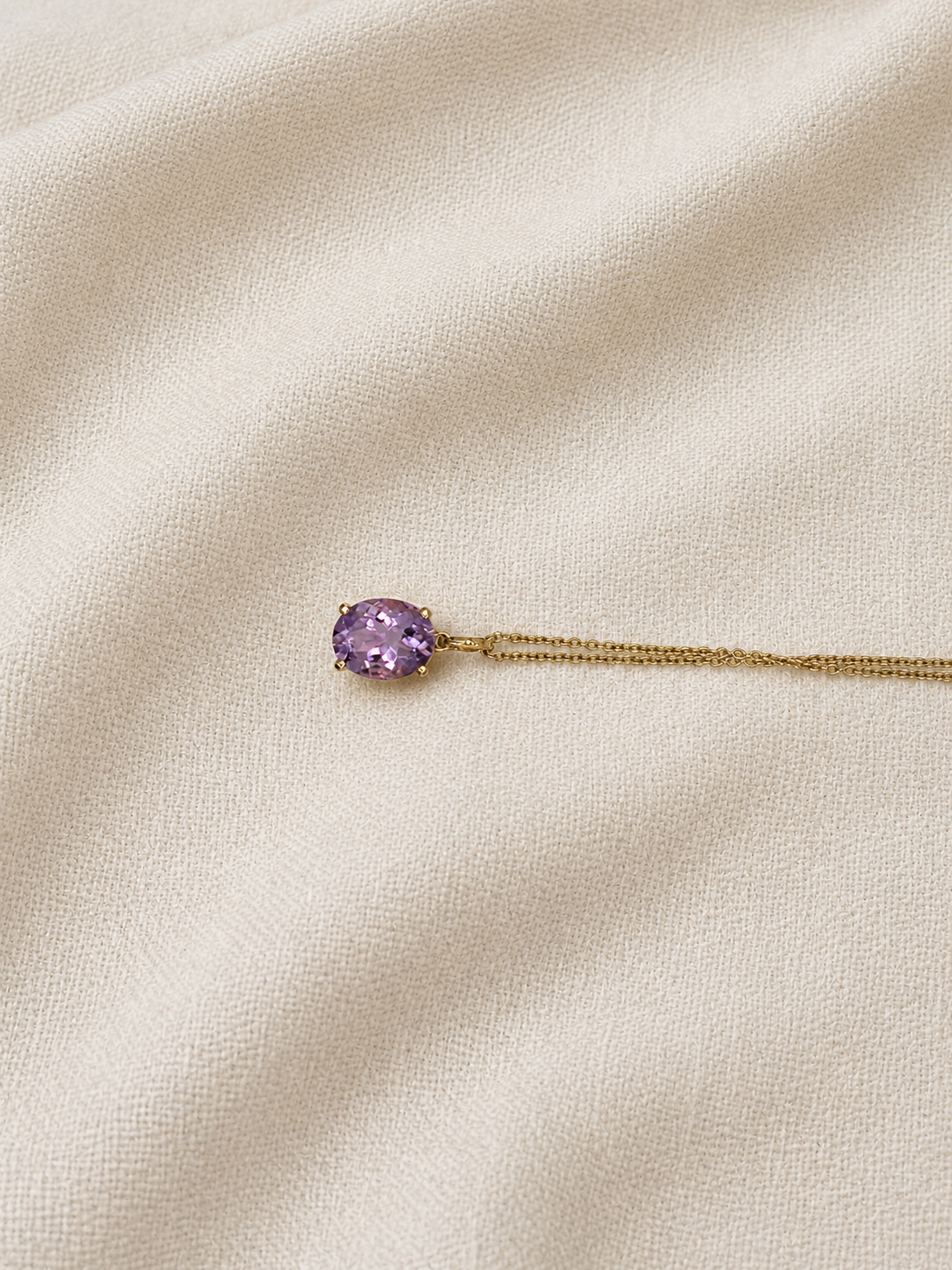 A solid 10k yellow gold chain necklace with an oval natural purple amethyst gemstone pendant by Ophelisse, displayed on a textured cream-colored fabric background.