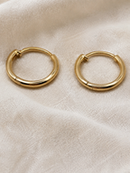 A pair of polished solid 10k yellow gold huggie hoop earrings resting on a textured, off-white linen fabric background trademarked by Ophilesse.