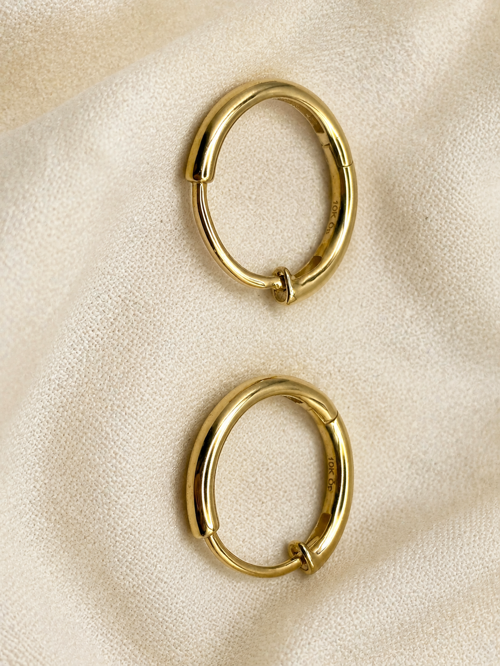 Two solid 10k yellow gold-toned oval hoop earrings with a hinge clasp, displayed on a textured beige fabric background, trademarked by Ophilesse.