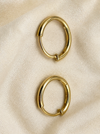 Two solid 10k yellow gold-toned oval hoop earrings with a hinge clasp, displayed on a textured beige fabric background, trademarked by Ophilesse.