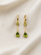 A pair of solid 10k yellow gold drop earrings with small round green peridot natural gemstones above larger pear-shaped natural green peridot gemstones, displayed on a textured off-white fabric background.