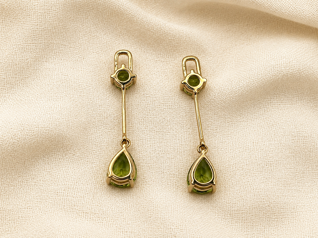 A pair of solid 10k yellow gold drop earrings with natural green peridot gemstones by Ophilesse, displayed on a textured beige fabric background.