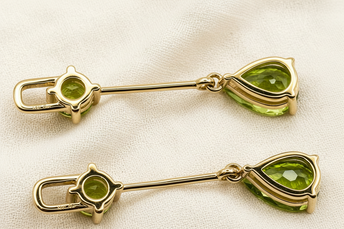 A pair of solid 10k yellow gold-toned lapel pins with round green peridot natural gemstones and teardrop-shaped green gemstones by Ophilesse, displayed on a textured beige fabric background.