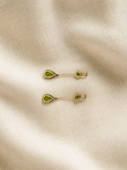 A pair of solid 10k yellow gold with green peridot teardrop-shaped stones and small round green stones, displayed on a textured beige fabric background.