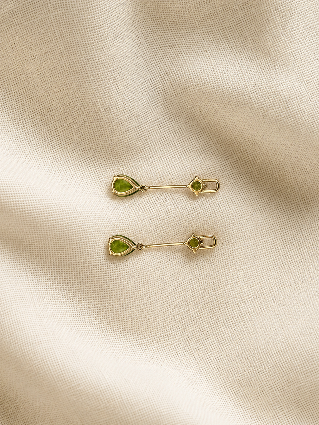 A pair of solid 10k yellow gold with green peridot teardrop-shaped stones and small round green stones, displayed on a textured beige fabric background.