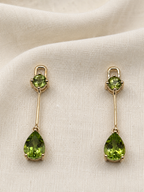 A pair of solid 10k yellow gold drop earrings with natural round green peridot gemstones at the top and teardrop-shaped natural green peridot gemstones at the bottom, displayed on a textured beige fabric background.