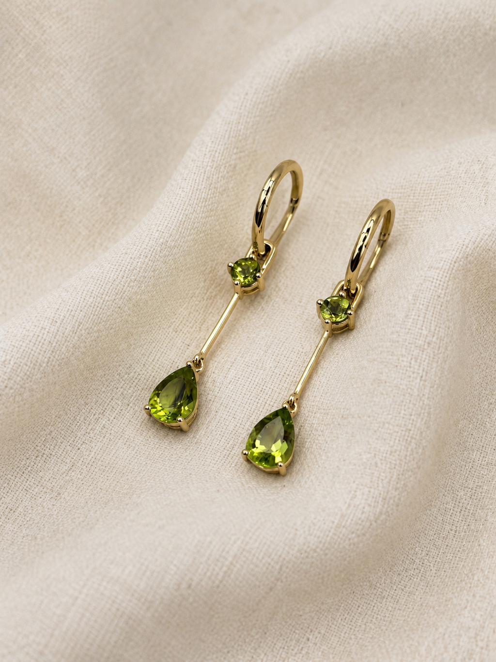 A pair of solid 10k yellow gold drop earrings with small round natural green peridot gemstones and larger teardrop-shaped natural green peridot gemstones, displayed on a textured beige fabric background.