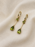 A pair of solid 10k yellow gold drop earrings with small round natural green peridot gemstones and larger teardrop-shaped natural green peridot gemstones, displayed on a textured beige fabric background.