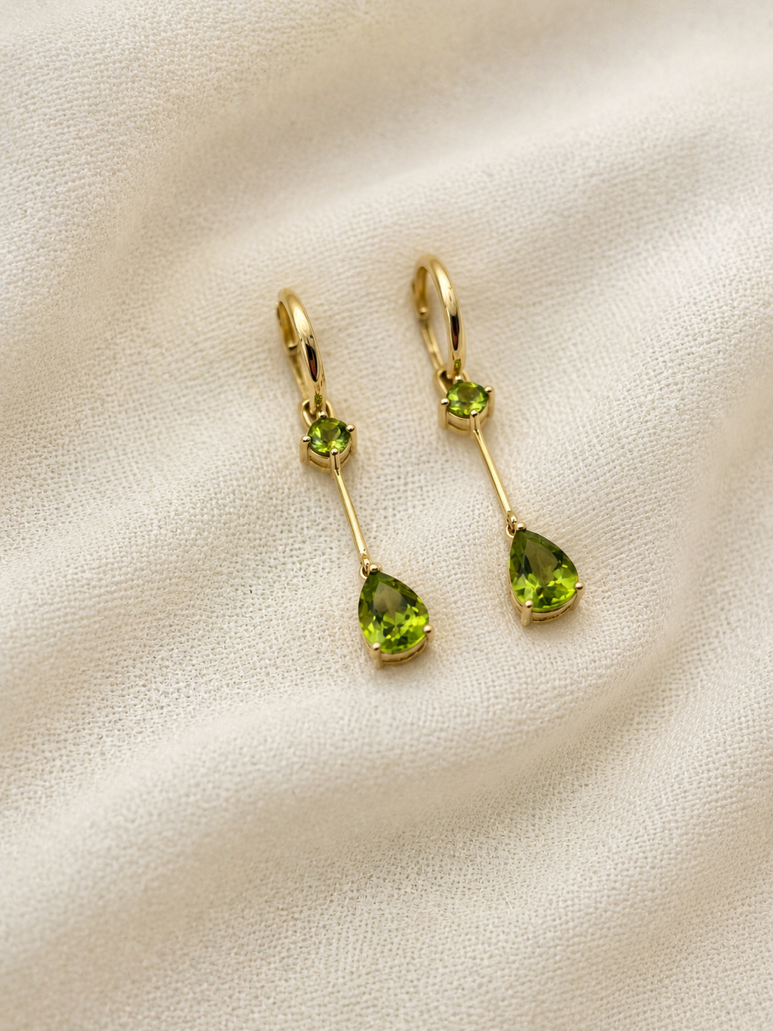 A pair of solid 10k yellow gold drop earrings with natural green peridot gemstones by Ophelisse, displayed on a textured beige fabric background.