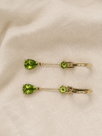 A pair of solid 10k yellow gold dangle earrings with natural green teardrop-shaped peridot gemstones and small natural round green peridot gems, displayed on a textured beige fabric background.