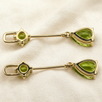 A pair of solid 10k yellow gold-toned lapel pins with round natural green peridot gemstones and teardrop-shaped green gemstones, displayed on a textured beige fabric background.