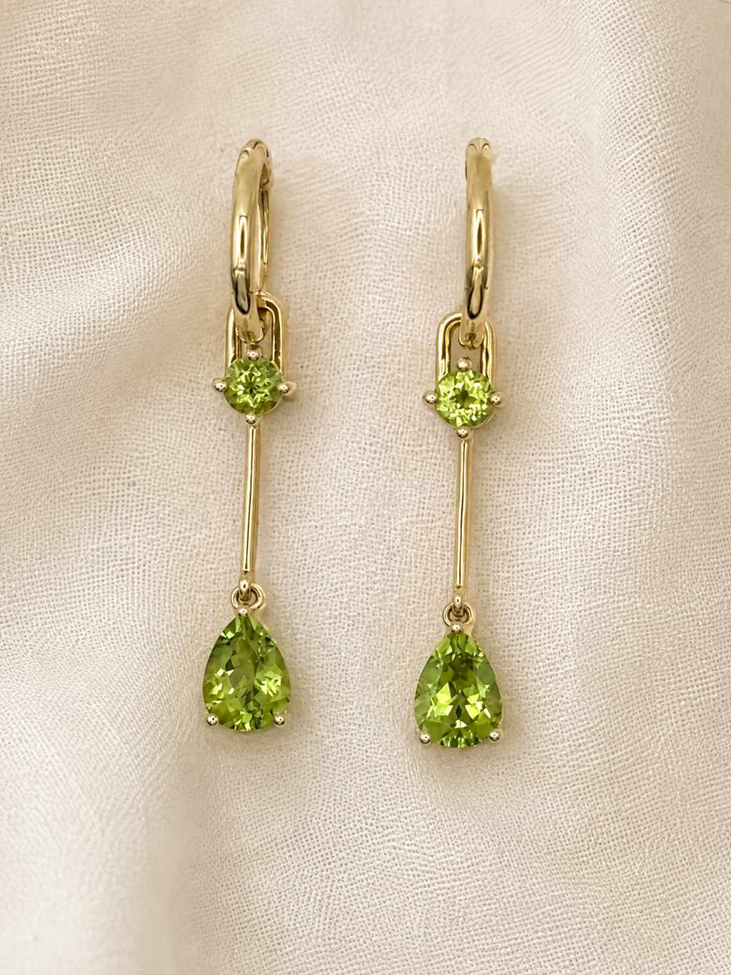 A pair of solid 10k yellow gold drop earrings with a small natural round green peridot gemstone above a larger pear-shaped natural green peridot gemstone by Ophelisse, displayed on a textured off-white fabric background.