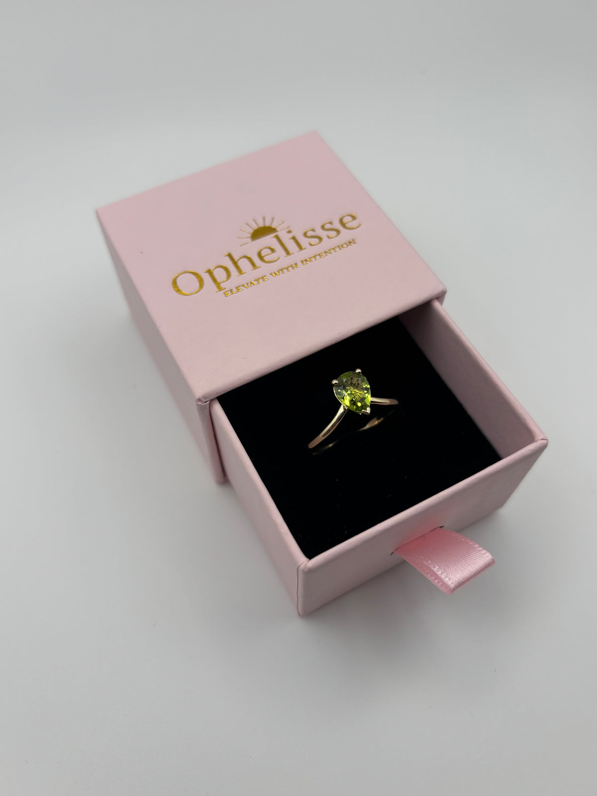 A yellow 10k gold luxurious ring with a natural green peridot gemstone in a gold setting, presented inside an open elegant pink Ophelisse jewelry box with a black velvet insert, on an light gray background.