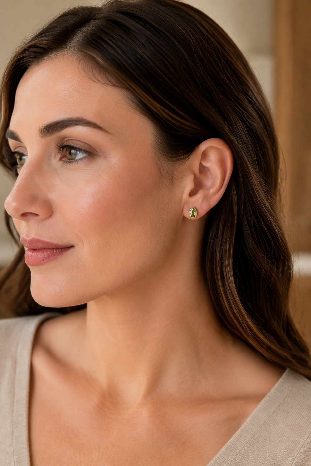 A close-up side profile of an elegant model with dark brown hair wearing a solid 10k yellow gold stud earring with a natural green peridot gemstone, set against a soft beige background.