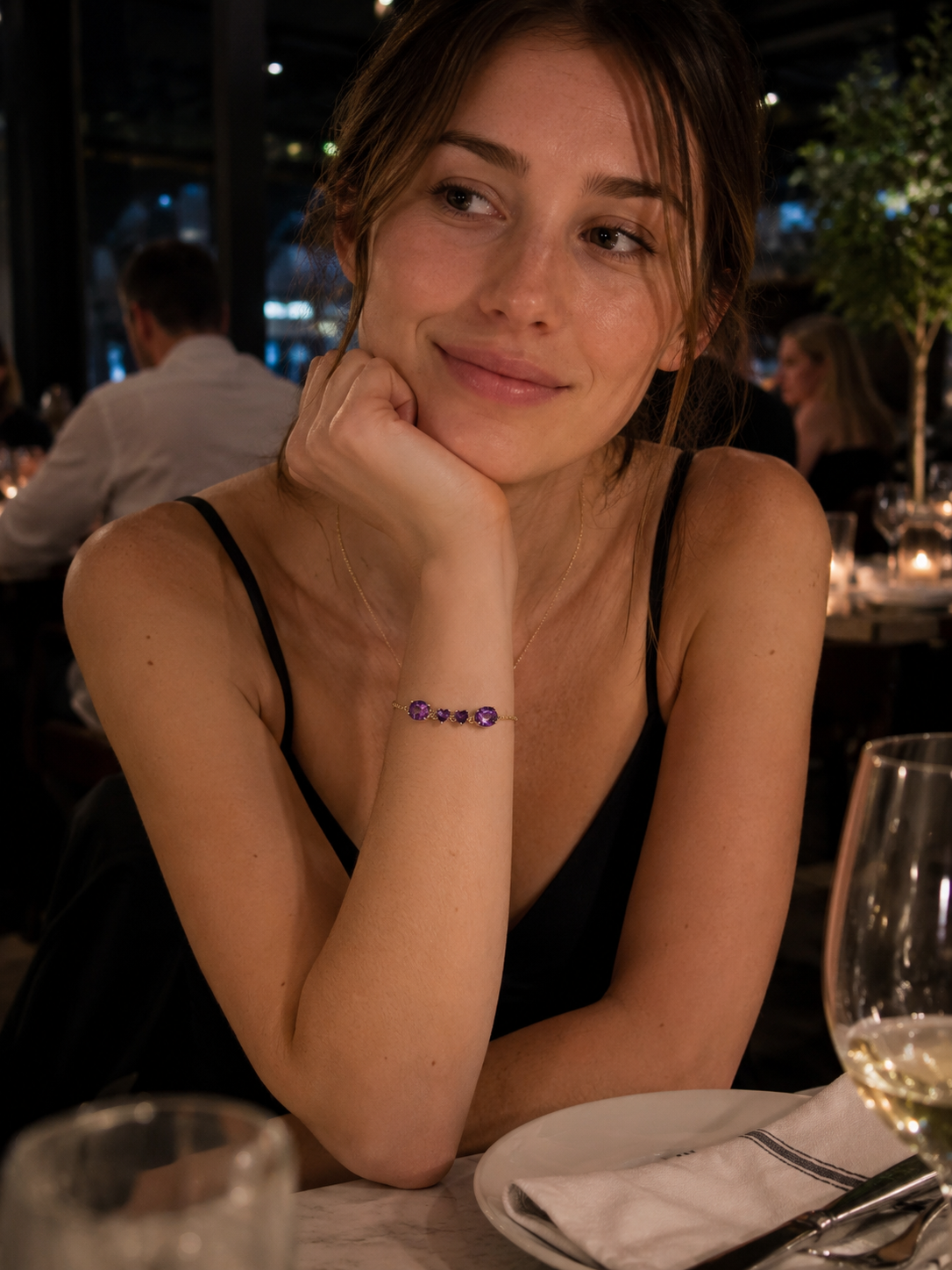 A woman wearing Ophelisse's bracelet on her wrist in a restaurant, the bracelet consists of solid 10k yellow gold with two large oval and two small heart natural purple amethyst gemstones.