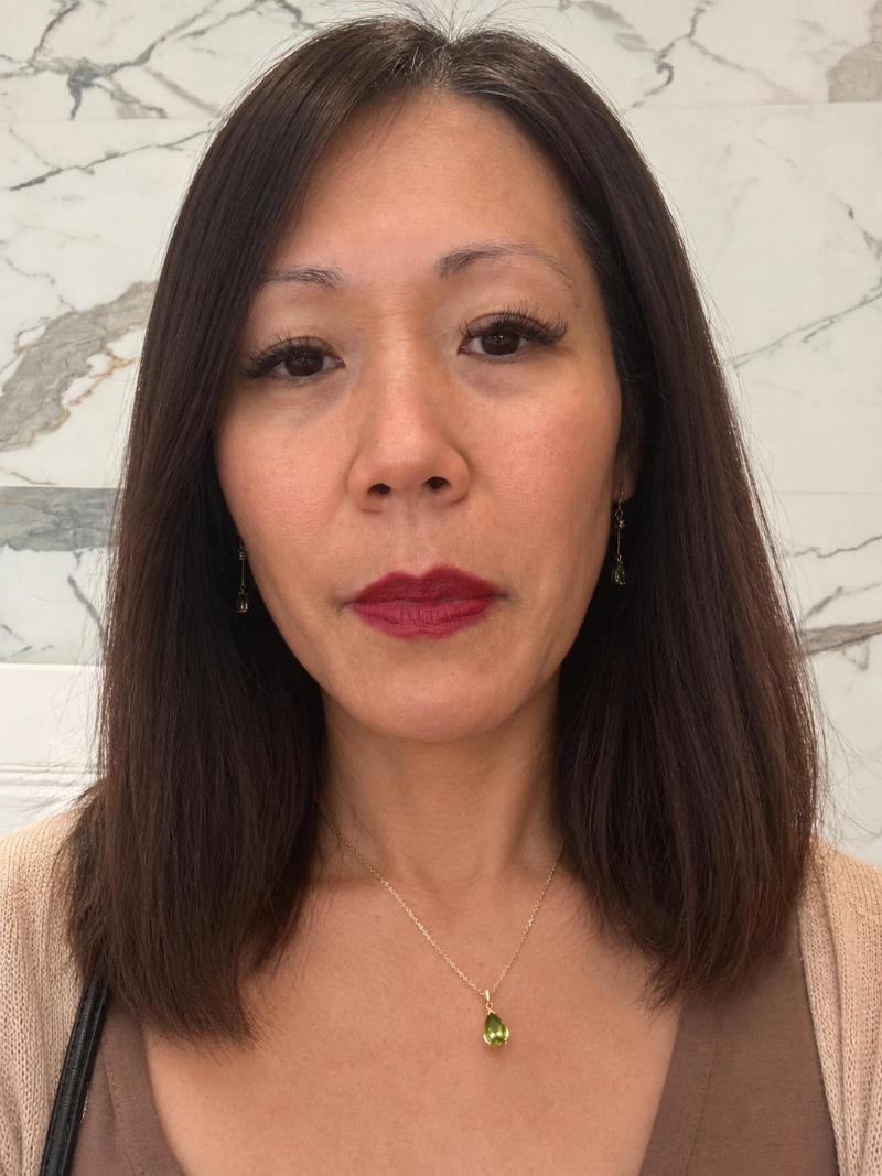 Ophelisse founder, Nha Clark, wearing 10k yellow gold necklace with a green peridot teardrop pendant, and peridot gold hoop earrings. She is standing in front of a light marble-patterned wall.