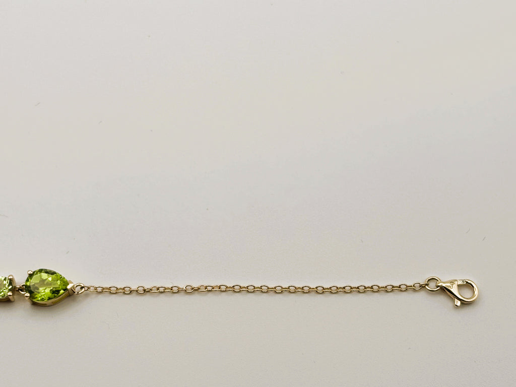 Picture displays part of Ophelisse's solid 10k gold chain bracelet with a pear-shaped natural green peridot gemstone and a smaller green stone, showing Ophelisse's trademark, shown against a plain light background.