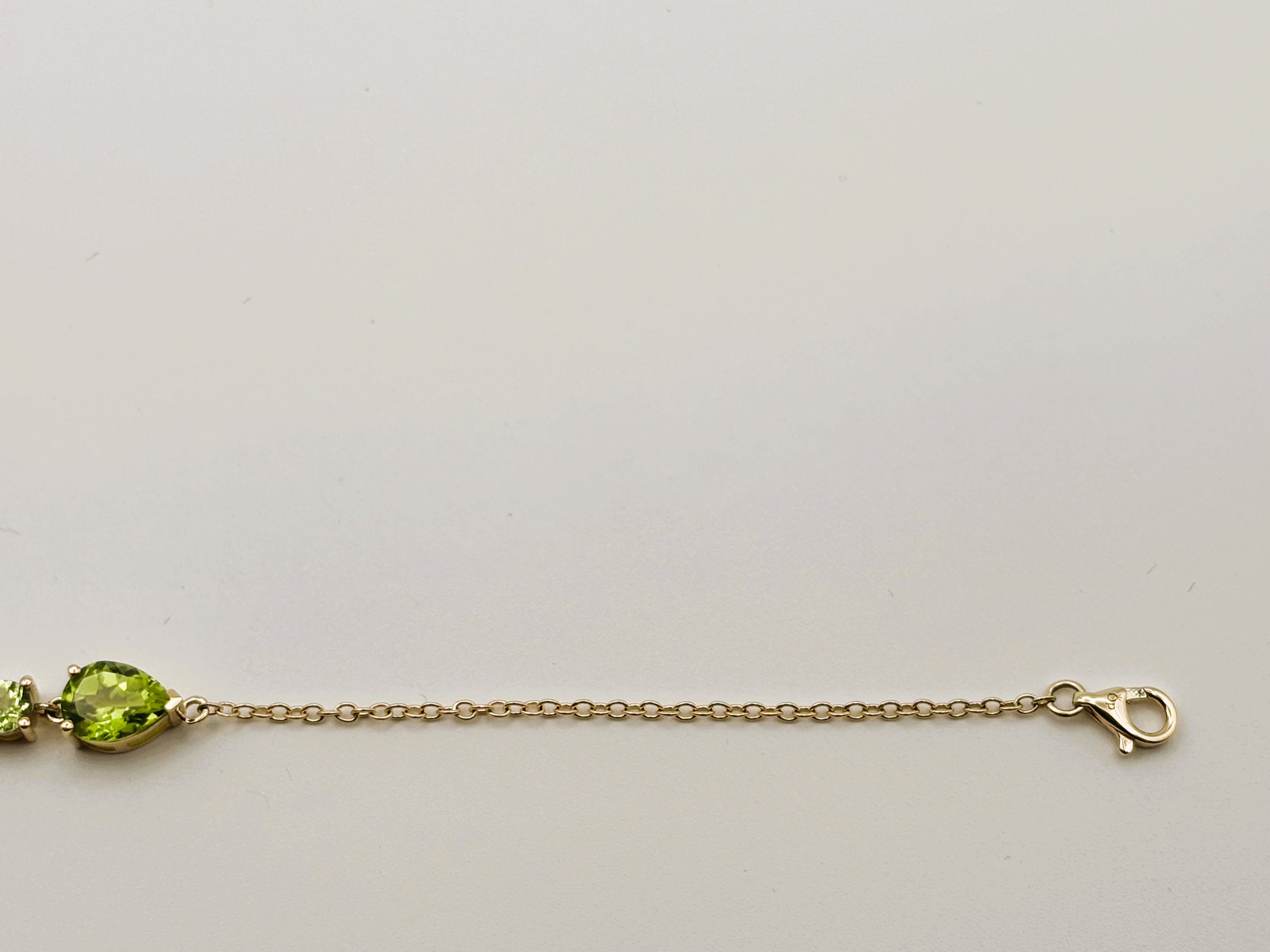 Picture displays part of Ophelisse's solid 10k gold chain bracelet with a pear-shaped natural green peridot gemstone and a smaller green stone, showing Ophelisse's trademark, shown against a plain light background.