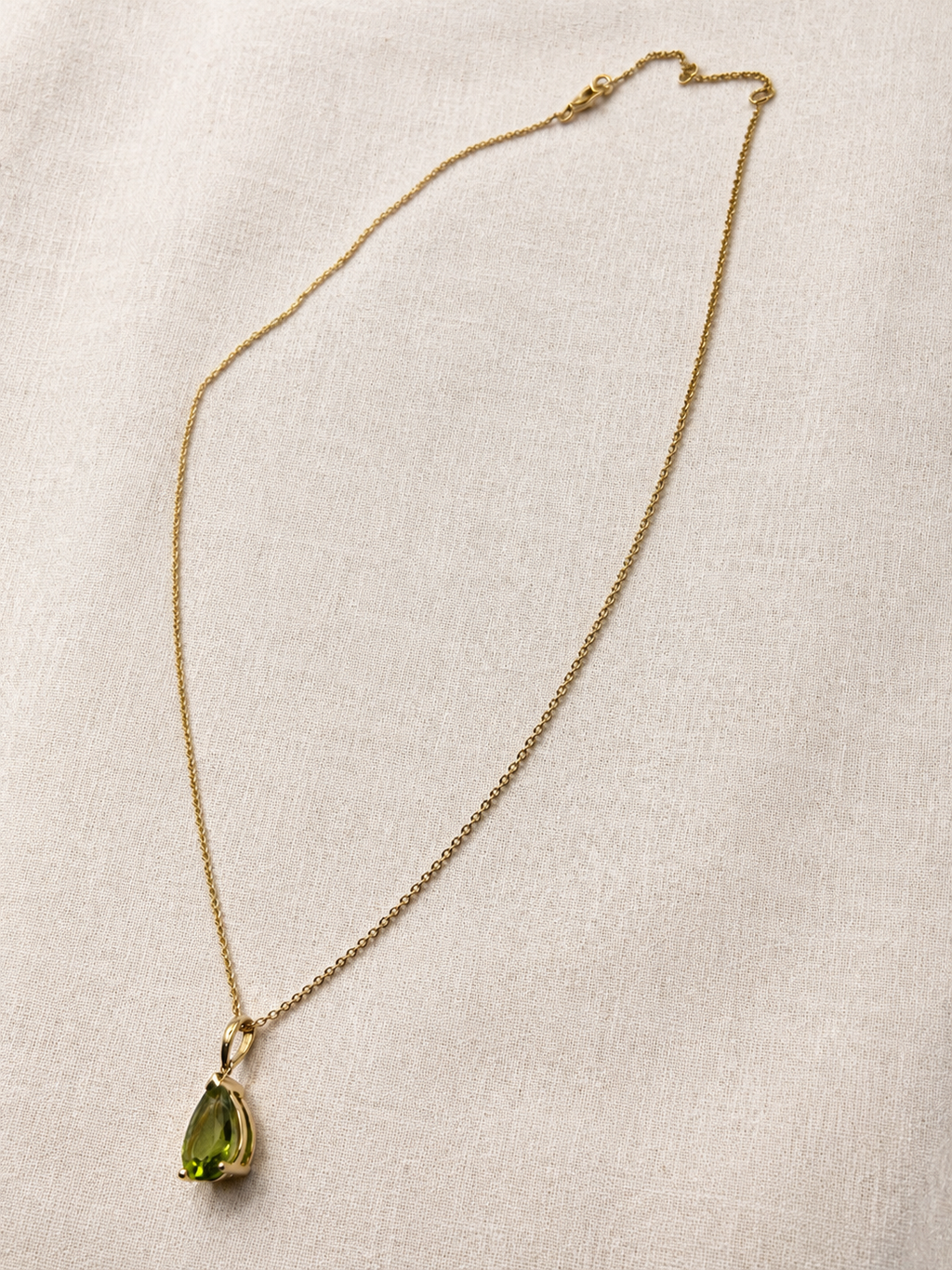 A delicate 10k yellow solid gold chain necklace with a teardrop-shaped luxurious green peridot gemstone pendant by Ophelisse, displayed on a luxurious textured off-white fabric background for elegant and quiet luxury.