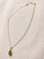 A delicate 10k yellow solid gold chain necklace with a teardrop-shaped luxurious green peridot gemstone pendant by Ophelisse, displayed on a luxurious textured off-white fabric background for elegant and quiet luxury.