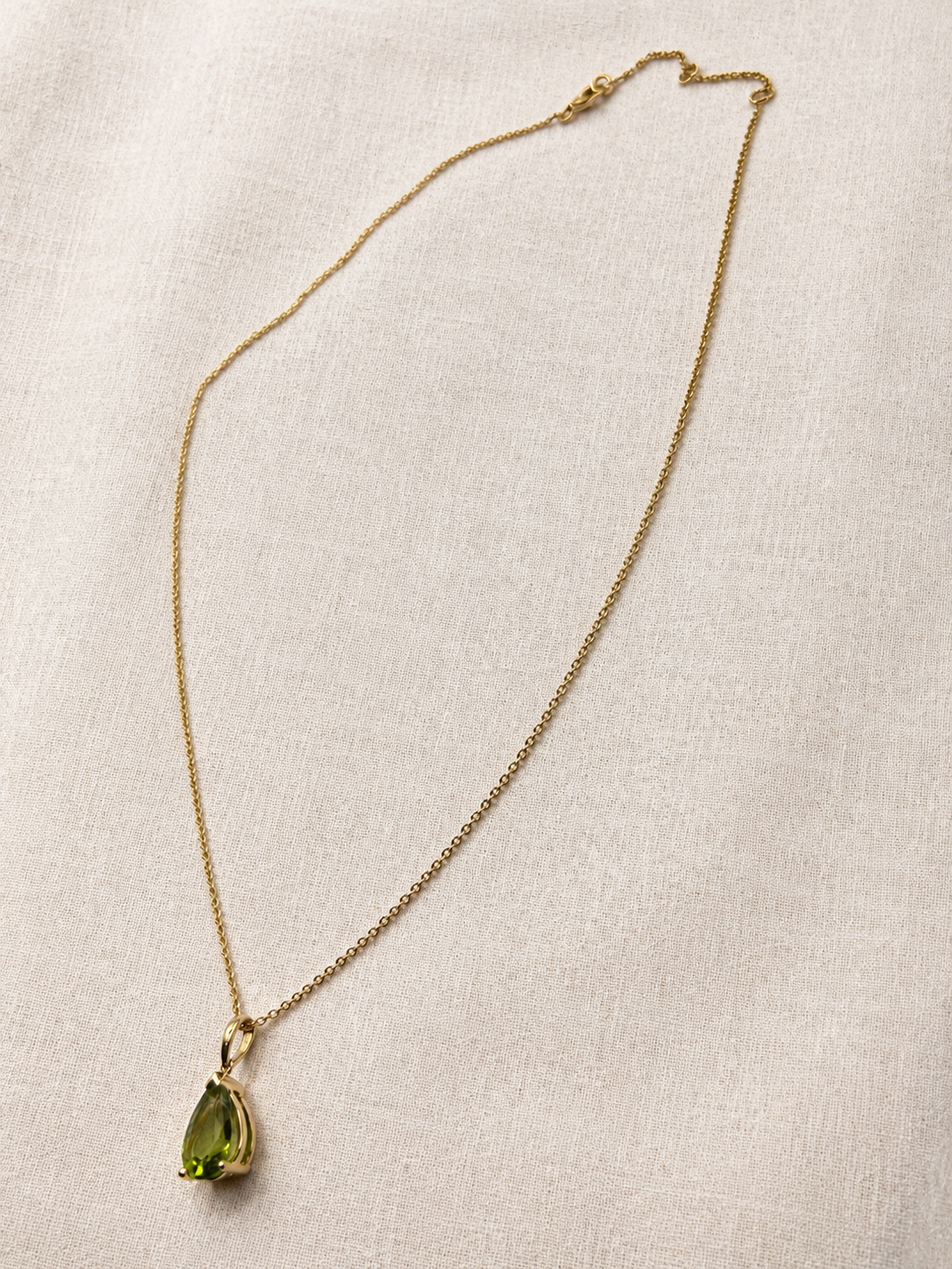 A delicate 10k yellow solid gold chain necklace with a teardrop-shaped luxurious green peridot gemstone pendant by Ophelisse, displayed on a luxurious textured off-white fabric background for elegant and quiet luxury.