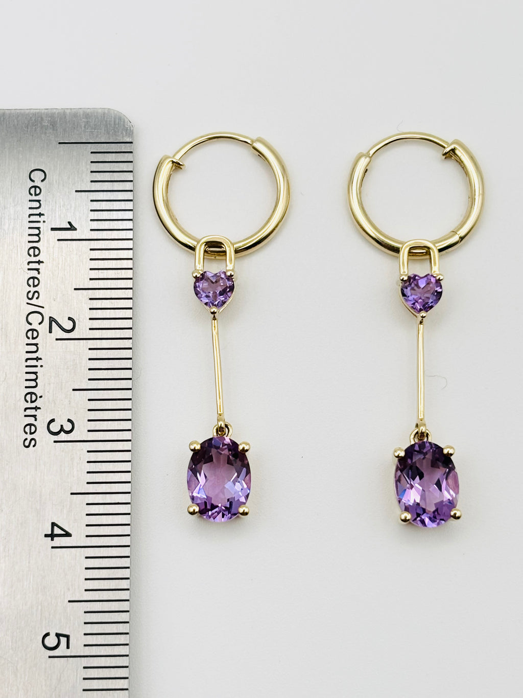 A pair of Ophelisse gold hoop drop earrings with small heart-shaped purple amethyst stones and larger oval purple amethyst drops, shown next to a ruler for measurement.