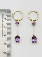A pair of Ophelisse gold hoop drop earrings with small heart-shaped purple amethyst stones and larger oval purple amethyst drops, shown next to a ruler for measurement.