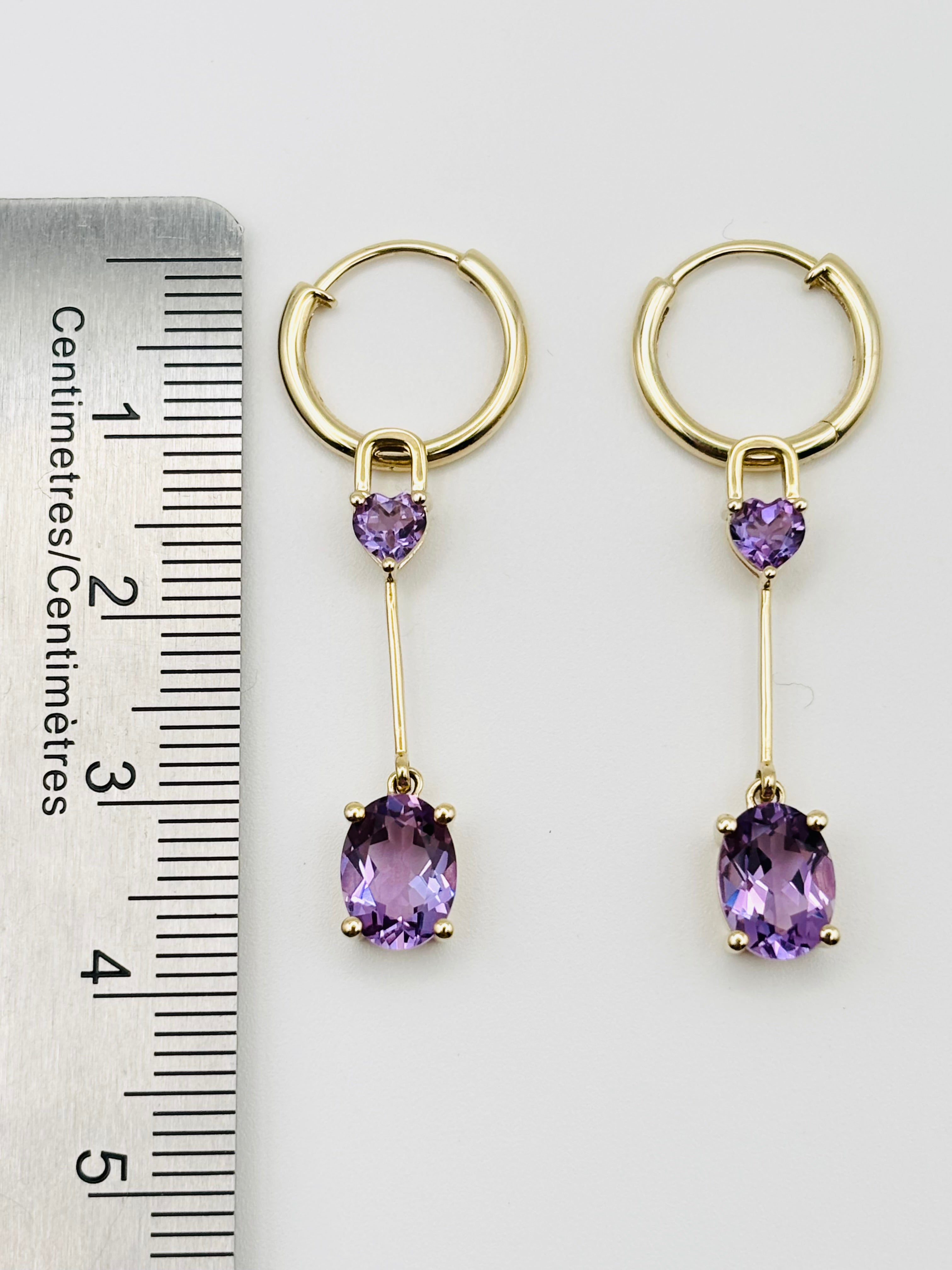 A pair of Ophelisse gold hoop drop earrings with small heart-shaped purple amethyst stones and larger oval purple amethyst drops, shown next to a ruler for measurement.
