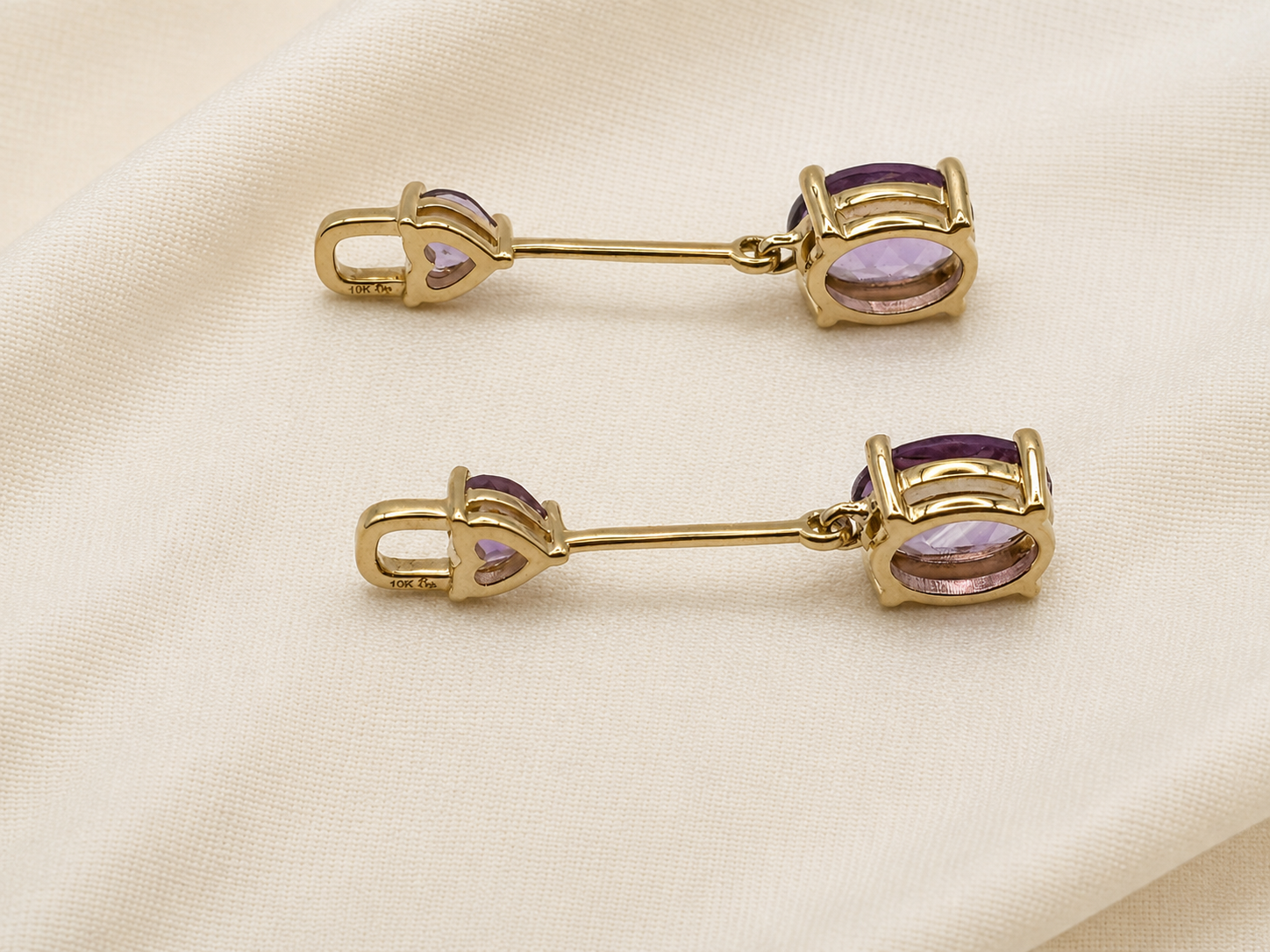 A pair of solid 10k yellow gold-toned bar pin brooches with oval purple natural amethyst stones by Ophelisse, displayed on a textured cream fabric background.